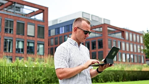 Man using a laptop and speaking into his hand outside office buildings during Stock Footage 348703121