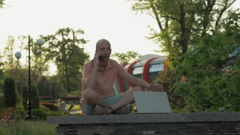 A man using a laptop and talking on the phone while sitting in a city park. Stock Footage 155497287