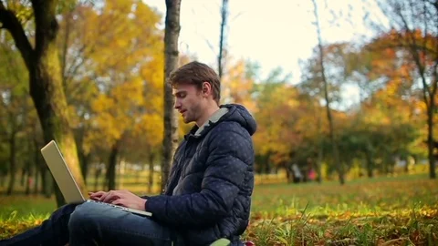 Man using laptop in autumn park working sitting on the grass, dolly camera Stock Footage 70682904