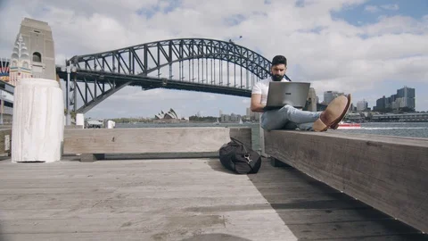 Man Using Laptop on Bench by Sydney Harbour Bridge. Low Angle Wide Shot Stock Footage 129363539