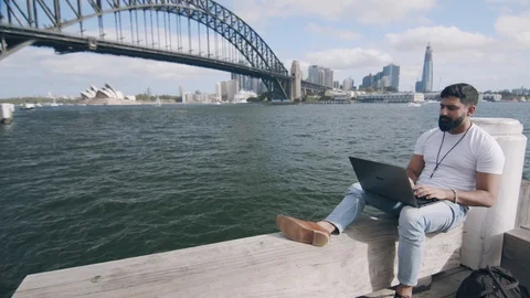 Man Using Laptop on Bench by Sydney Harbour Bridge. Wide Circle Shot Stock Footage 129363636