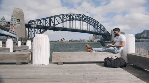 Man Using Laptop on Bench by Sydney Harbour Bridge. Static Wide Shot Stock Footage 129363855