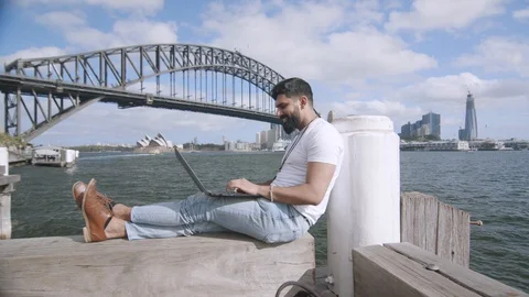 Man Using Laptop on Bench by Sydney Harbour Bridge. Wide Dolly Left Stock Footage 129486032