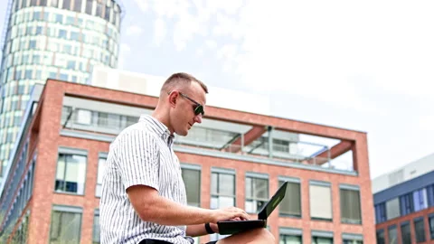 Man using a laptop outside a brick office building in an urban business district Stock Footage 348704000