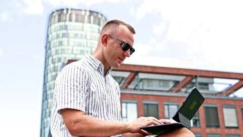 Man using a laptop outside a brick office building beside a glass tower during Stock Footage 348704253