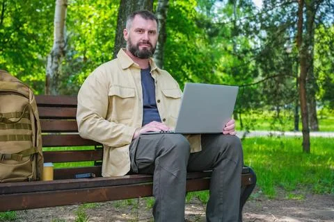 Man using laptop on park bench with backpack and coffee, concept of remote .. Foto stock