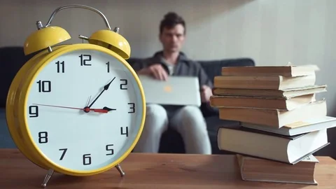 Man using laptop sitting on couch reading book at home. Yellow clock is ticking Stock Footage 80814633