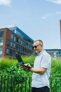 Man using laptop in urban area with green plants and buildings around him d.. Stock Photos