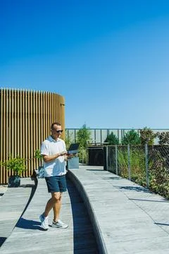 Man using laptop while walking on wooden pathway near a modern building on .. Stock Photos
