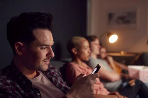 Man Using Mobile Phone Whilst Friends Watch TV At Home In Evening Stock Photos