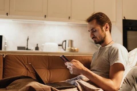 Man Using Phone on Sofa with Laptop Nearby: Relaxed Evening in a Cozy Kitch.. Stock Photos