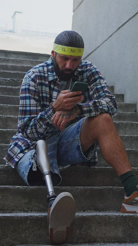 Man using a phone while sitting on steps with a prosthetic leg in an urban area Stock Footage 291961920