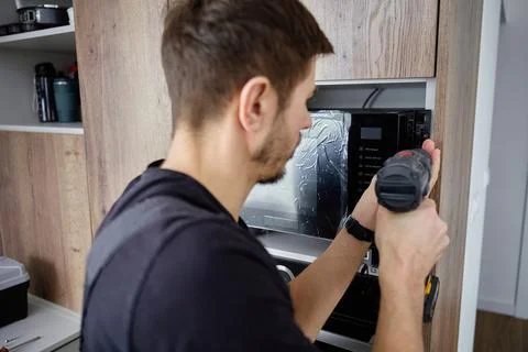 Man Using Power Drill to Fix Microwave Oven in Kitchen Cabinet Stock Photos