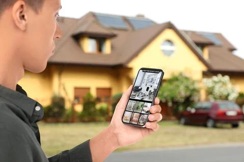 Man using smart home security system on smartphone near house outdoors. Dev.. Stock Photos