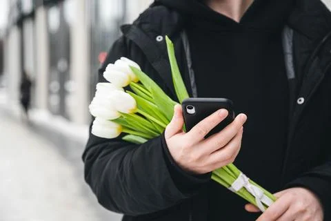 A man is using a smartphone and holding a bouquet of white tulips outside. Stock Photos
