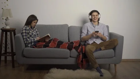 Man using smartphone and listening music while girlfriend reading book on sofa 스톡 동영상 124705049
