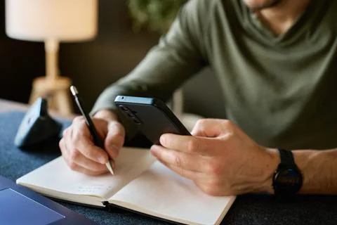Man using smartphone and writing notes at desk Stock Photos