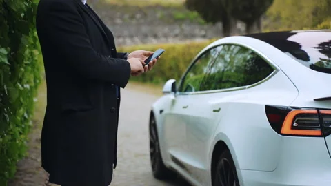 Man using smartphone app for remote controlling of white premium class Tesla car 스톡 동영상 163445539