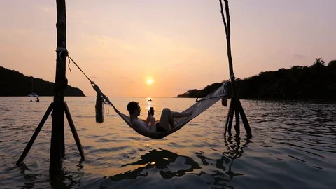 Man using smartphone on the beach hammock at sunset, mobile phone and travel. Video stock 103125110