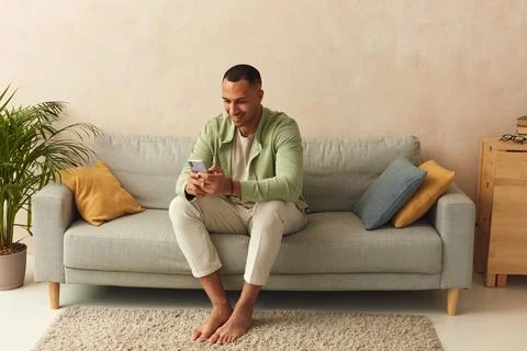 Man Using Smartphone At Home. Handsome Multiracial Guy Sitting On Sofa Stockfoto's