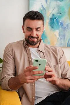 Man using smartphone indoors smiling at screen while seated on yellow chair Fotos Stock