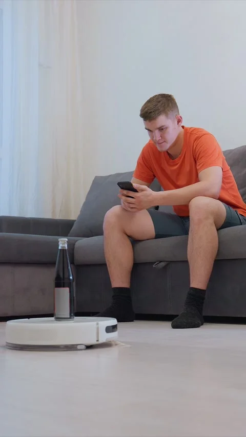 Man Using Smartphone Observing Robotic Vacuum Cleaner in Modern Living Room Stock Footage 346795846
