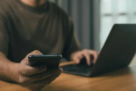 Man using smartphone in one hand while typing on laptop at wooden table wit.. Foto stock