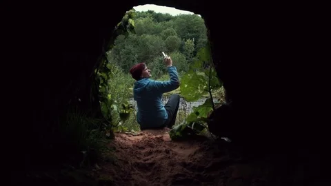 Man using smartphone sitting near exit from cave. Deep forest at background. Stock Footage 80053825