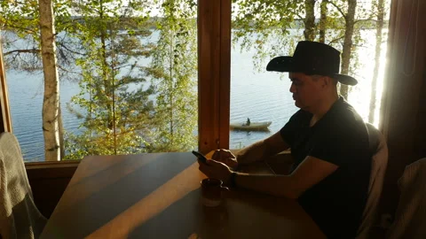 Man using smartphone sitting at table by window of cottage and waving hand hello Stock Footage 95430426