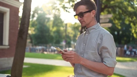 Man using smartphone standing outdoors. Bright sun and green trees at background Video stock 79357337