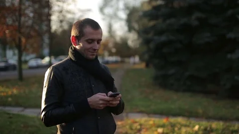 Man using smartphone typing and chatting, smiling outdoors in autumn city street Stock Footage 71252330