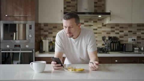 Man using smartphone while having breakfast in kitchen. A man in a t-shirt .. Stock Footage 332287866