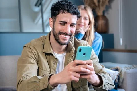 Man using a smartphone while sitting at home with a woman in the background. Stock Photos