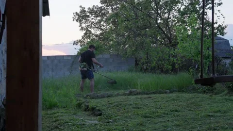 Man Using String Trimmer to Cut Tall Grass in Rustic Backyard at Dusk Stock Footage 341767011