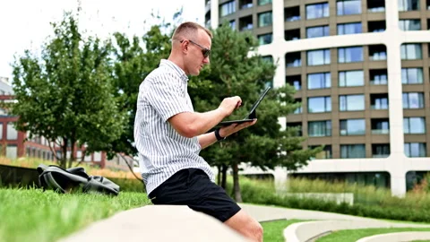 Man using a tablet while seated on a low wall beside grass and office buildings Stock Footage 348700635