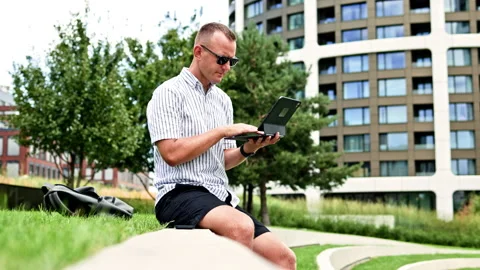 Man using a tablet while seated on a concrete ledge outside an office building Stock Footage 348702062