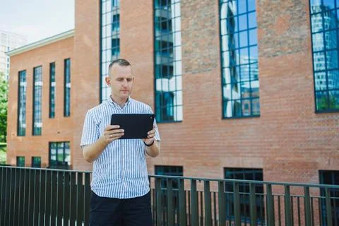 Man using a tablet while standing outside near a modern brick building on a.. 스톡 사진