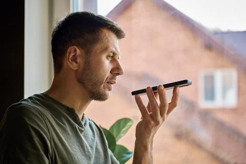 Man using voice assistant on smartphone by window at home Stock Photos
