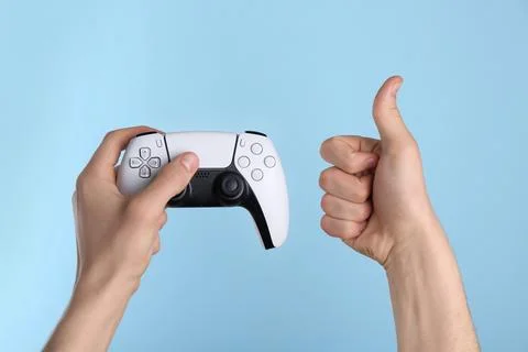 Man using wireless game controller and showing thumbs up on light blue back.. Stock Photos