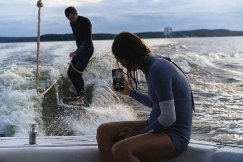 Man wake surfing on lake while woman films him during sunset Photos