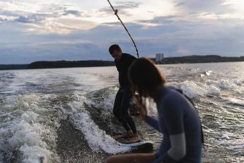 Man wake surfing on lake while woman records him at sunset Photos