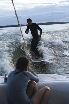 Man wake surfing on lake while woman films him during sunset Photos