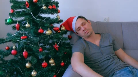Man is waking up sitting on couch at home near Christmas tree at eve, hangover. Stock Footage 136519994