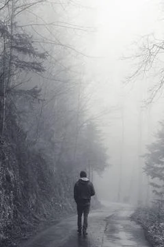Man walking alone on a road through a dark foggy forest Stock Photos