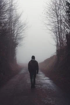 Man walking alone on a road through a dark foggy forest Stock Photos