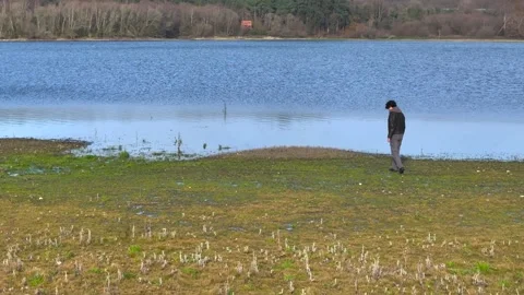 A Man Is Walking Alone Towards Calm Lake During Daytime. Aerial Drone Shot Video stock 265441766