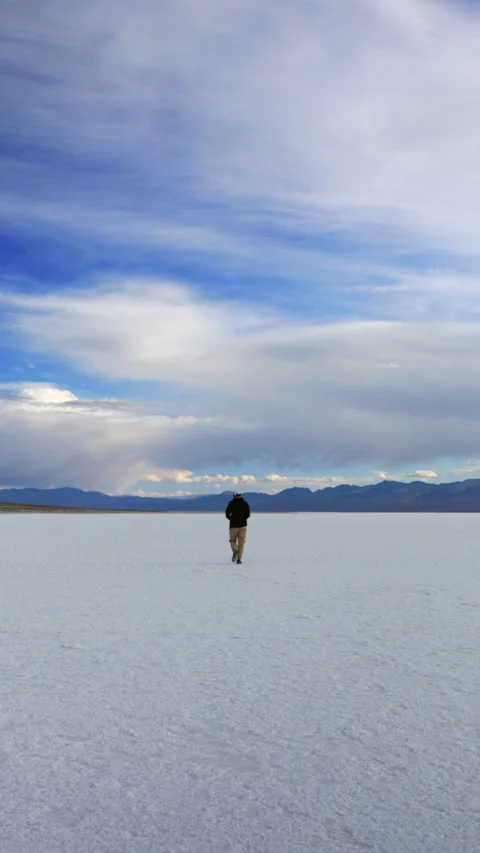 Man Walking Alone on Vast Salt Flats Under Dramatic Blue Sky and Mountains. Vídeo Stock 342918085
