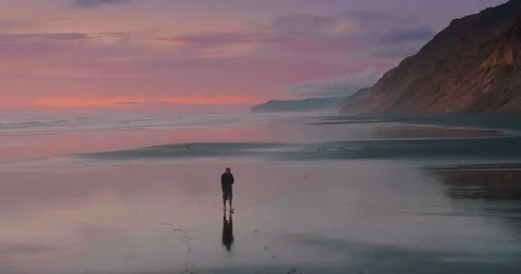 Man walking along the beach at low tide with the shore reflecting the sky giving Stock Footage 279612849