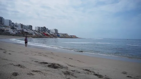 A man walking along the seashore with the sand, waves and buildings at Senoritas 库存影片 195126133