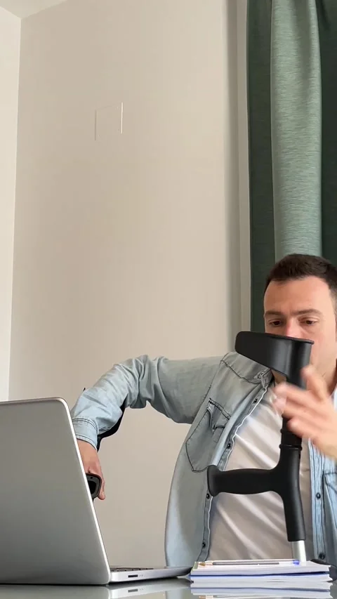 Man walking on crutches sits in front of laptop to work from home or have an Stock Footage 236062726